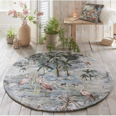 A round rug with a tropical pattern featuring flamingos and palm trees, placed on a wooden floor in a room with decorative pillows, a vase with flowers, and a bench.
